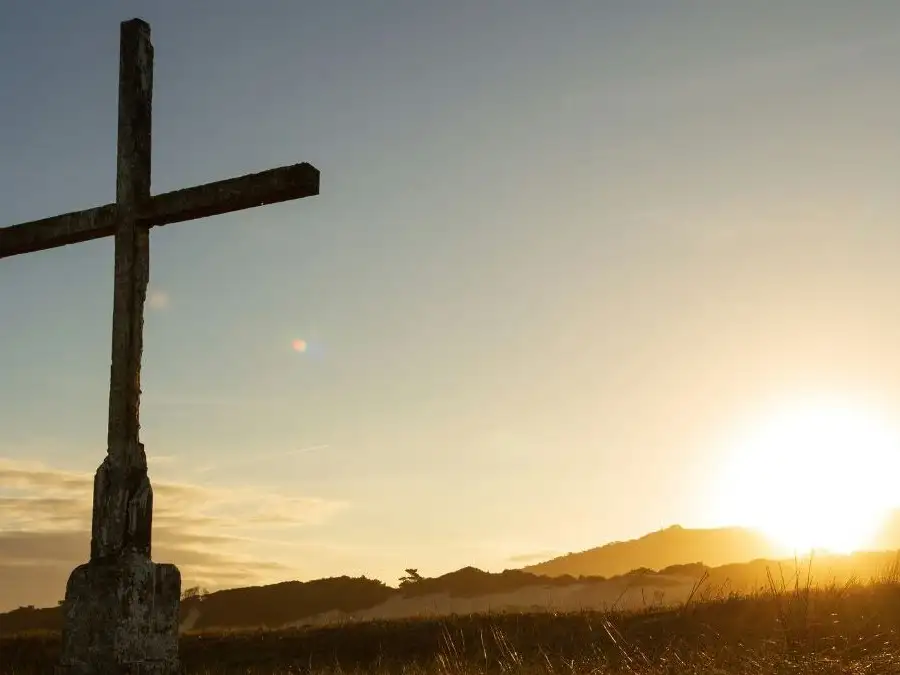 A large cross in a field in the foreground, with a the sun setting behind.